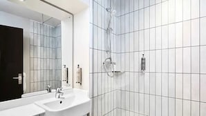 A modern bathroom with white tiled walls, a large mirror, two sinks, and a shower. - Premier Inn Cambridge City Centre (Cambridge)