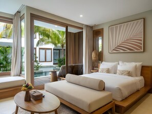 A modern hotel room with a large bed, a sofa, a small round table, and a view of a pool and greenery. - Cocana Resort (Gili Trawangan)