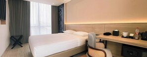 Habitación de hotel con cama, escritorio con silla, cafetera, una botella, y una mesita con taza y caja de pañuelos.