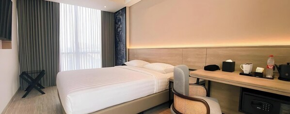 Habitación de hotel con cama, escritorio con silla, cafetera, una botella, y una mesita con taza y caja de pañuelos.