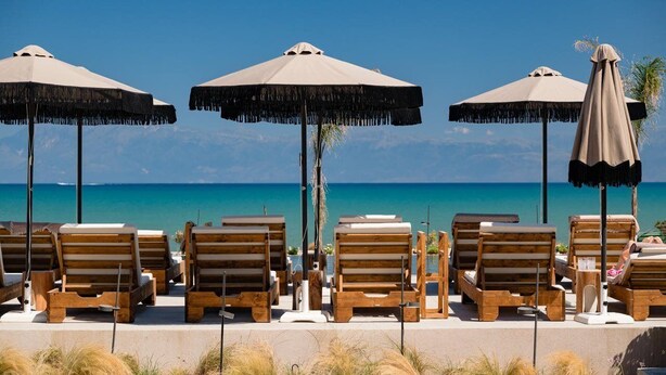 Ein Strandloungebereich mit Holzliegen und Reetdachschirmen mit Blick auf das klare, blaue Meer.