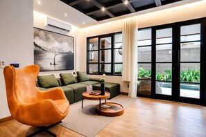 Ein modernes Wohnzimmer mit einer Couch, einem einzigartigen orangefarbenen Sessel, einem kleinen runden Tisch und einem großen Fenster mit Blick auf Grünflächen.