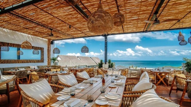 An outdoor dining area with wicker furniture, a wooden table set for a meal, and a view of the ocean. - Secana Beachtown (Canggu)