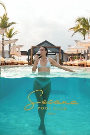 Una mujer con traje de baño blanco en una piscina del Secana Pool Club en Bali, con sillones de descanso y sombrillas al fondo.