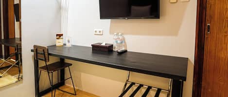 Una habitación con escritorio, silla, televisión montada en la pared, una mesita con caja de pañuelos y botellas de agua, zapatero y puerta de madera.