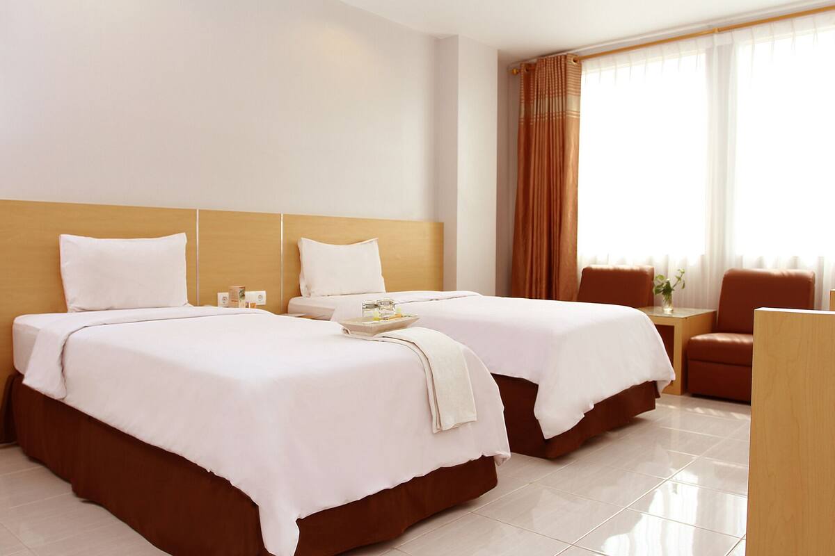 Een hotelkamer met twee bedden, een houten hoofdbord, een tafeltje, een stoel en een raam met gordijnen.