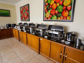 A buffet setup with multiple serving stations, a coffee machine, and a large fruit painting on the wall. - Hotel Grande Lampung Powered by Archipelago (Bandar Lampung)