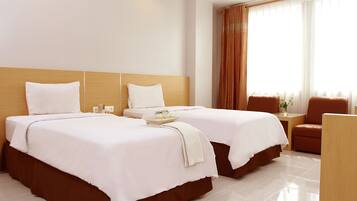 A hotel room with two beds, a wooden headboard, a small table, a chair, and a window with curtains.