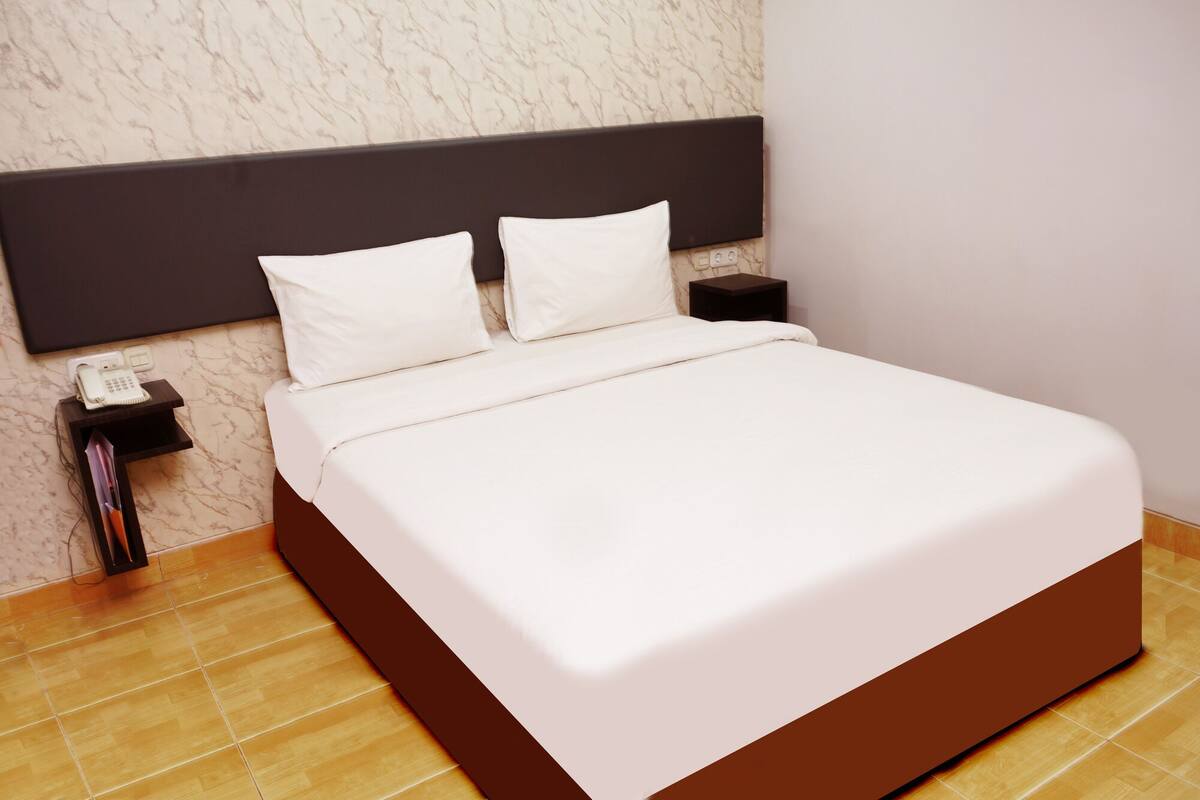 Een opgemaakt bed met een donker hoofdbord, wit beddengoed en een klein nachtkastje met een telefoon en een lamp.
