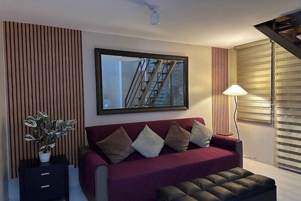 A modern living room with a red sofa, a black ottoman, a potted plant, a framed mirror, and a standing lamp. - PRS at One Palm Tree Villa Near Airport (Pasay)
