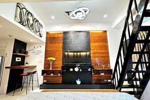 A modern living room with a large flat-screen TV, wooden paneling, a black fireplace, and a staircase. - PRS at One Palm Tree Villa Near Airport (Pasay)
