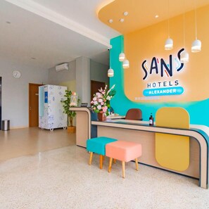 A reception desk with a sign for Sans Hotels Alexander, a clock, and a refrigerator in the background. - Sans Hotel Alexander Bandung (Bandung)