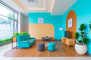 A modern lobby with a teal sofa, two ottomans, a wooden cabinet, and a large potted plant. - Sans Hotel Alexander Bandung (Bandung)
