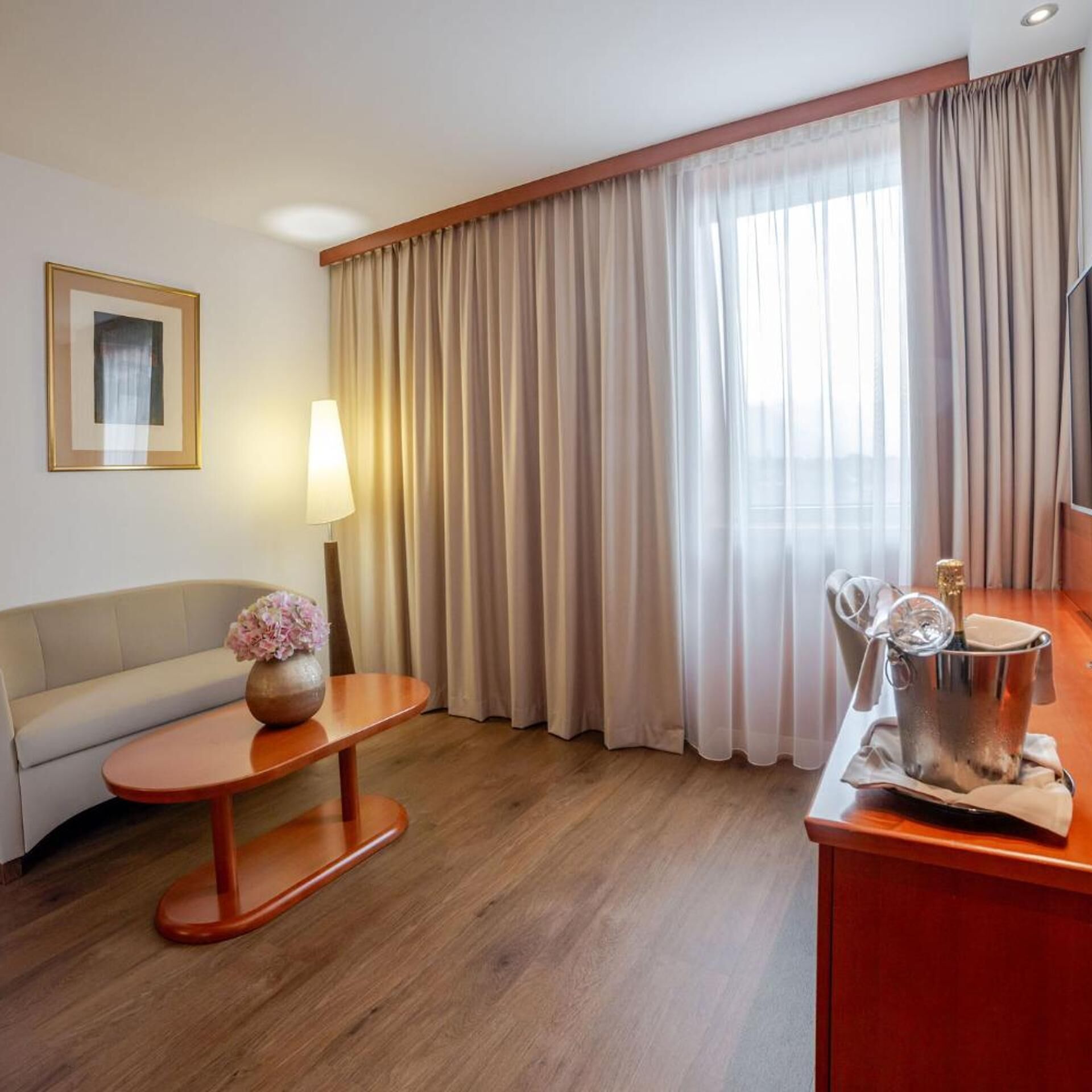 Ein Hotelzimmer mit einer Couch, einem kleinen Tisch mit Blumenvase, einer Lampe, einem Fernseher und einer Minibar mit einer Champagnerflasche.