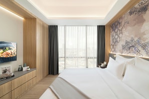 A hotel room with a bed, a TV, a desk with a magazine, and a view of the city through large windows.