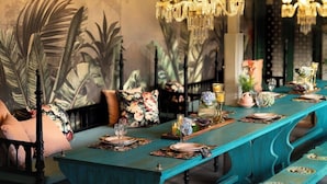 Une longue table à manger recouverte d’une nappe turquoise, mise avec des assiettes, des verres et des objets décoratifs.