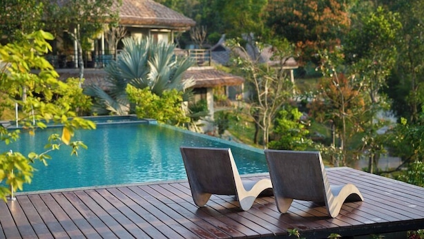 Une terrasse en bois avec deux chaises longues donnant sur une piscine, le tout entouré d’une végétation luxuriante et d’un bâtiment traditionnel doté d’un toit de chaume.