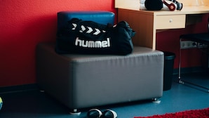 Una habitación pequeña con una pared roja, un escritorio de madera, una bolsa negra con la etiqueta "hummel", un par de zapatos negros y una alfombra roja.