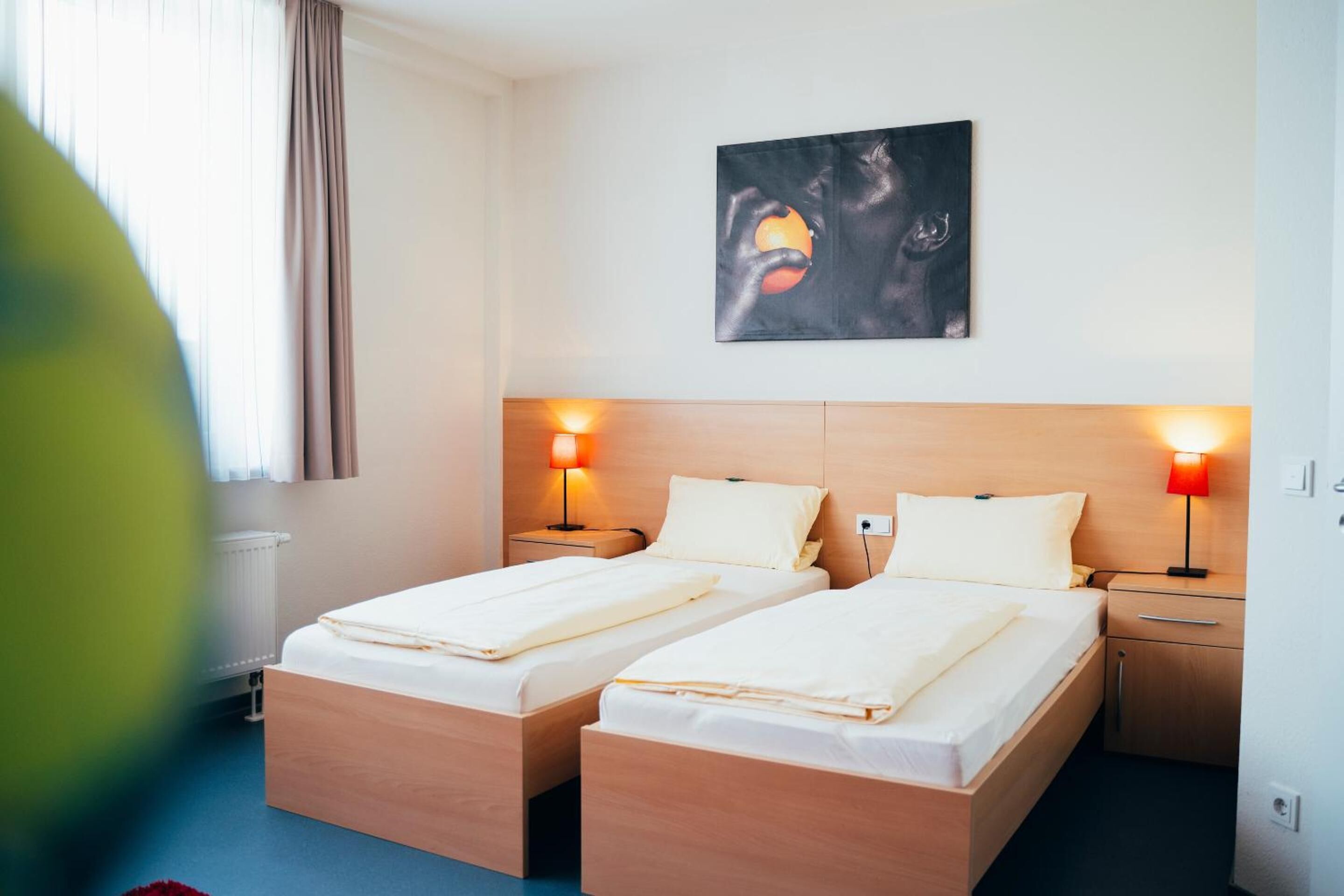 A hotel room with two single beds, a nightstand with a lamp, a wall-mounted artwork, and a window with curtains.