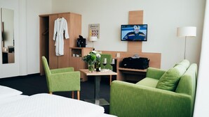 Habitación de hotel con cama, sillas verdes, sofá, televisor, mesa con jarra y copas de vino, y armario con bata.