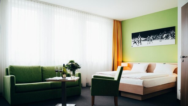 Ein Hotelzimmer mit einem grünen Sofa, einem kleinen Tisch mit Weingläsern, einem Sessel, einem Bett und einem an der Wand hängenden Bild.