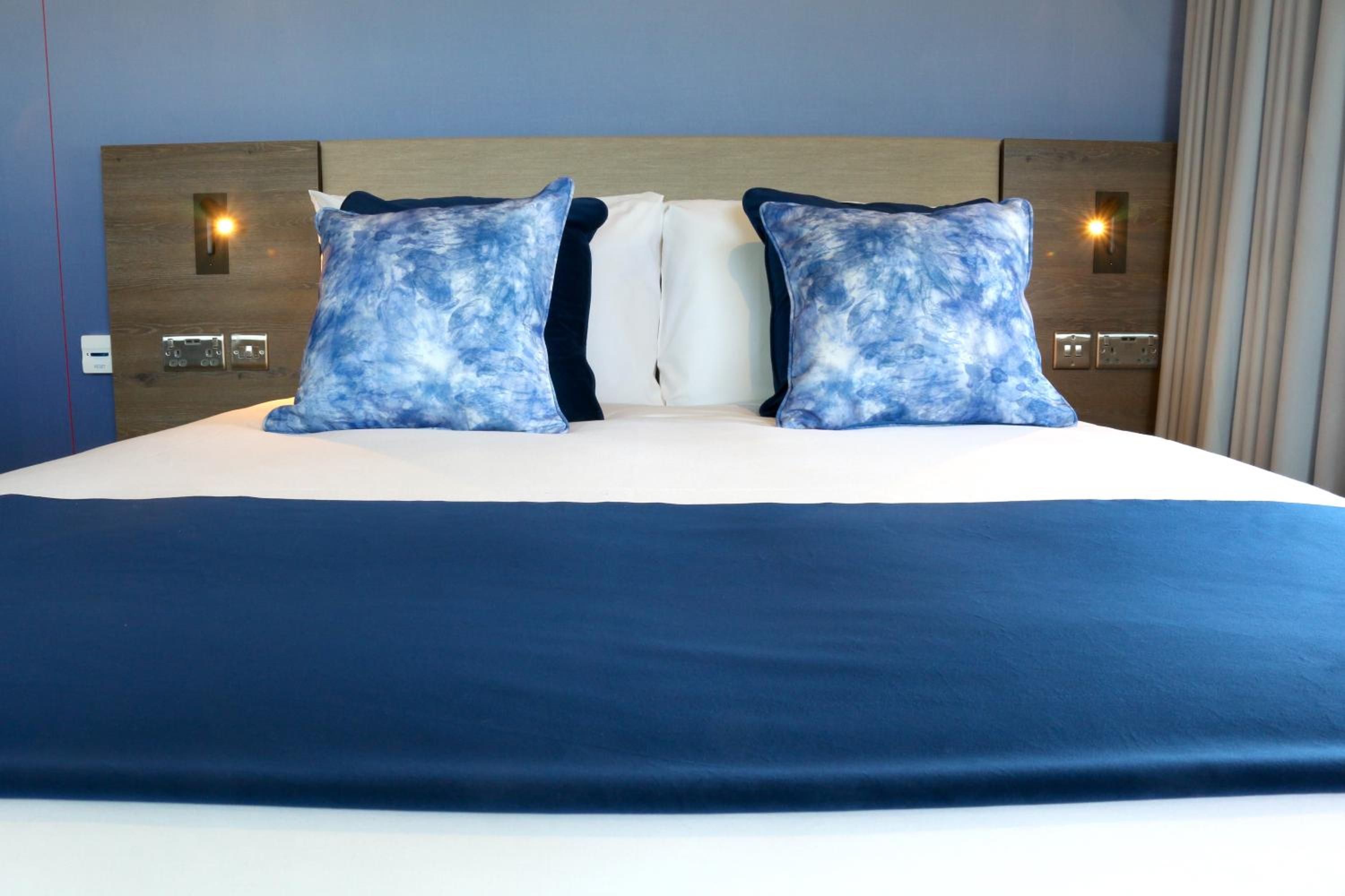A hotel room with a large bed, two pillows, a wooden headboard, and wall-mounted light fixtures.