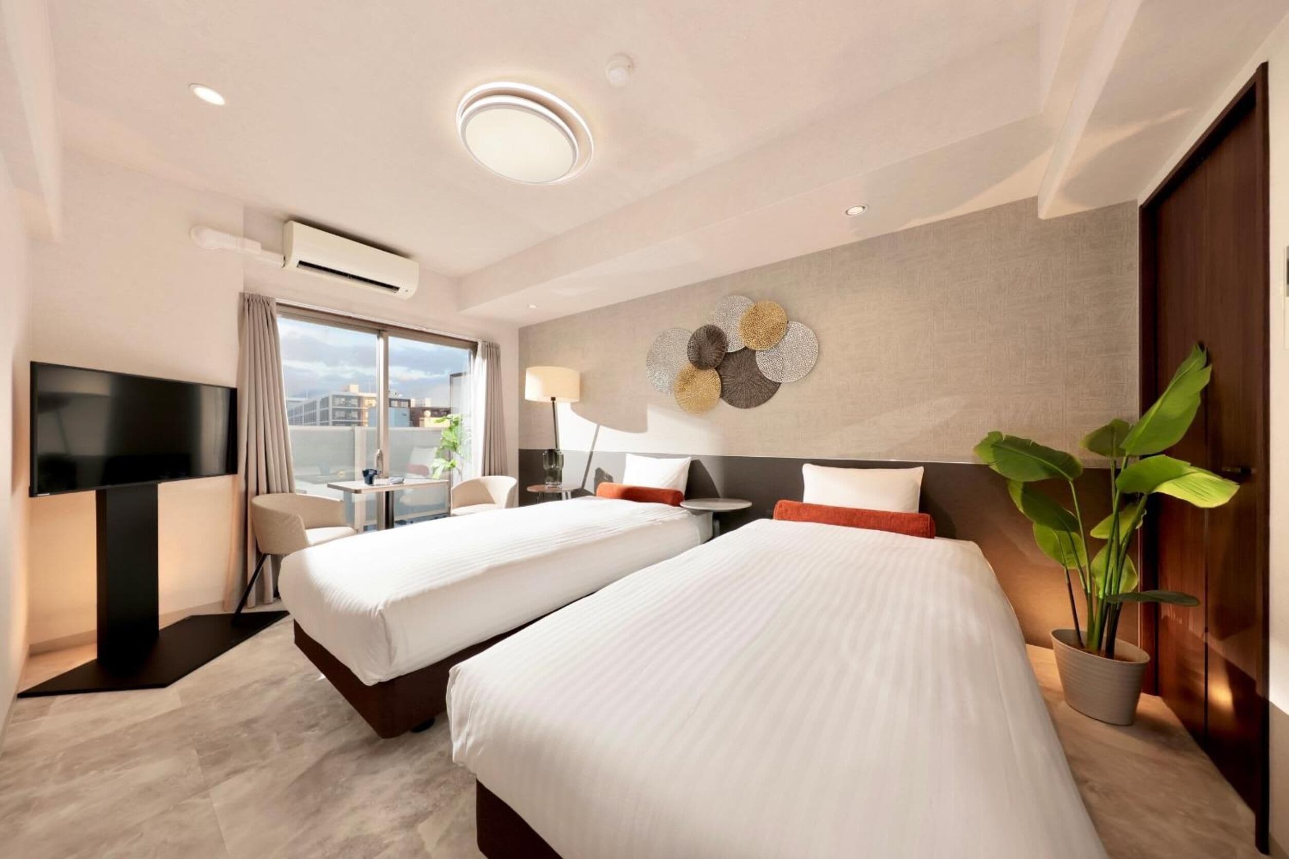 A hotel room with two beds, a TV, a desk, a lamp, and a potted plant.