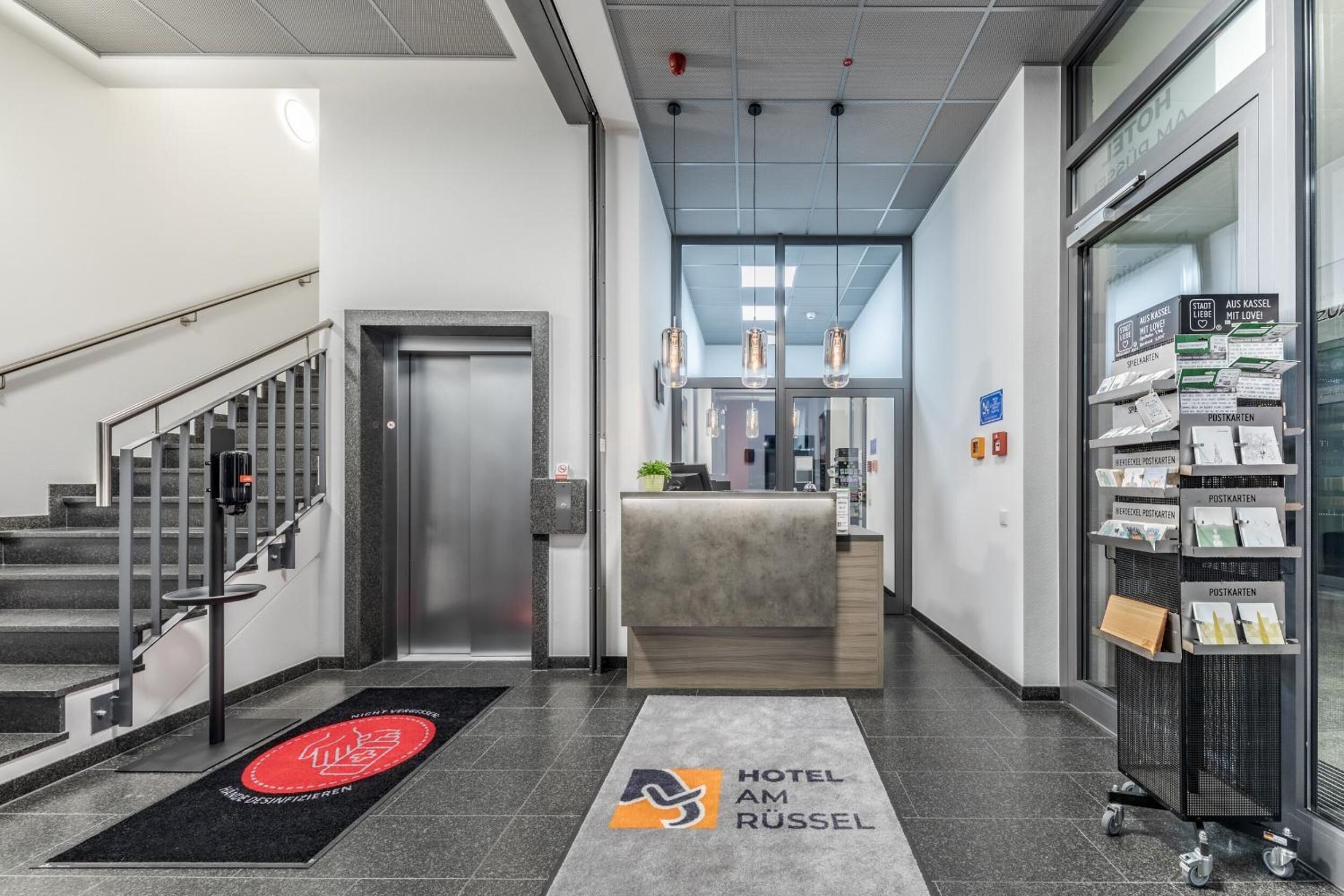 Eine Hotelhalle mit einer Rezeption, einer Treppe und einer Postkartenpräsentation.