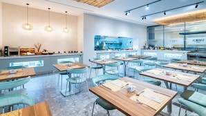 Un ristorante moderno con tavoli di legno e sedie color petrolio, un'area cucina e lampade a sospensione.