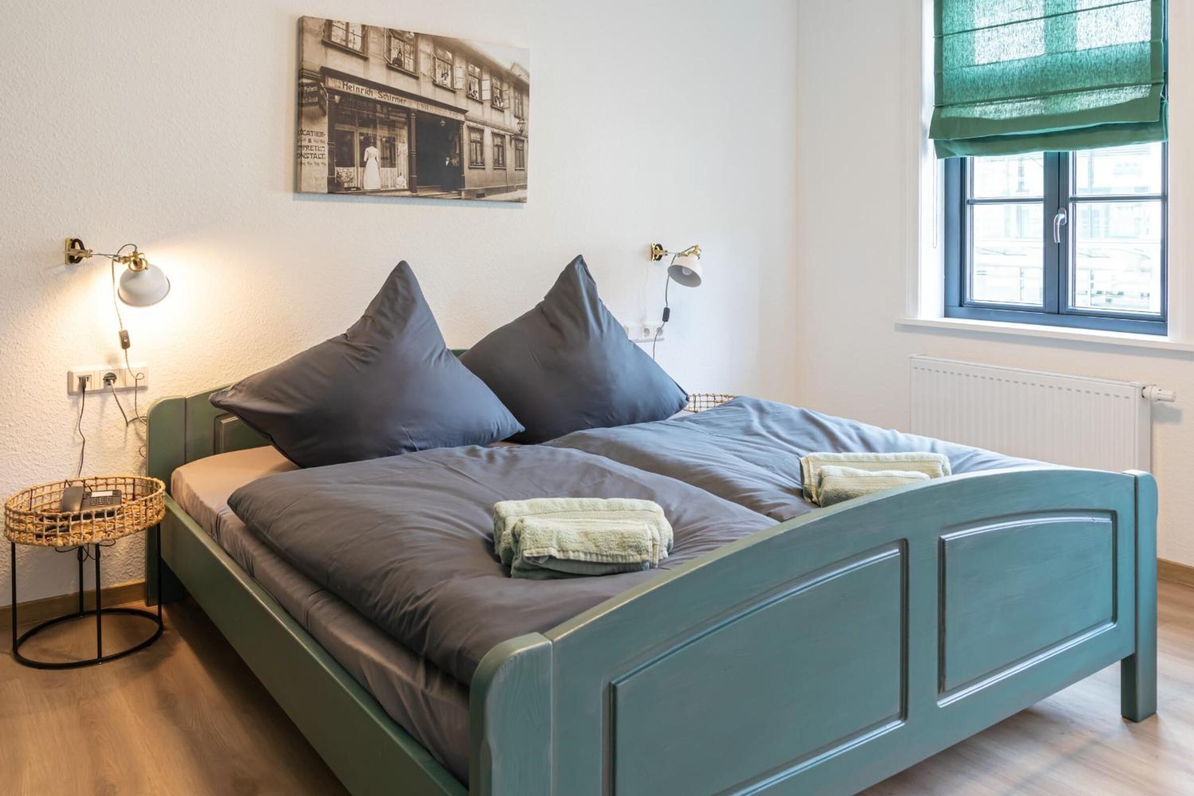 Un dormitorio con una cama, dos lámparas de noche, una ventana con persiana verde, un radiador y un cuadro colgado en la pared.