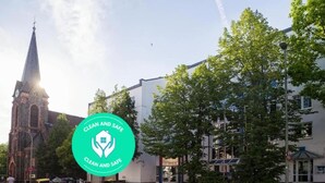 A church with a tall steeple, a modern building with a white facade, and a green sign with 'CLEAN AND SAFE' text. - Hotel B99 (Offenbach am Main)