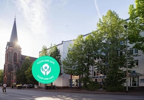 A church with a tall steeple, a modern building with a white facade, and a green sign with 'CLEAN AND SAFE' text. - Hotel B99 (Offenbach am Main)
