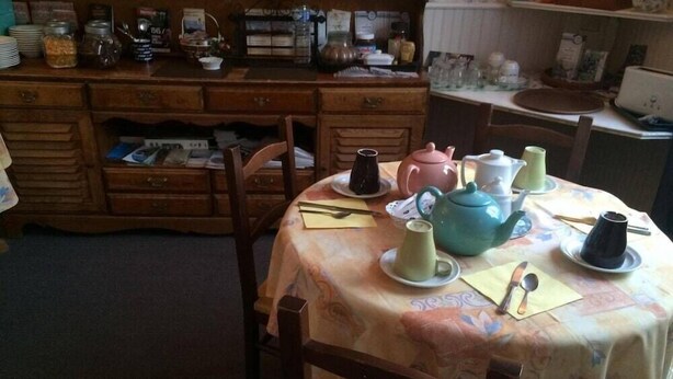 A dining table with a floral tablecloth, surrounded by wooden chairs. There are various teapots and cups on the table. - Hotel Bernieres (Caen)