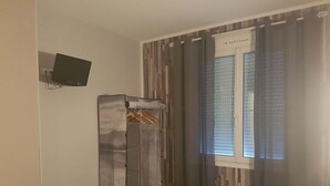 A bedroom with a bed, a wardrobe, a TV mounted on the wall, and a window with blinds. - Hotel Bernieres (Caen)