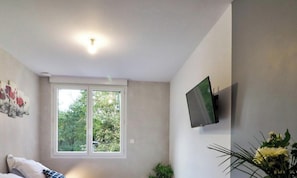 A room with a bed, a TV mounted on the wall, a window with a view of trees, and a ceiling light. - Appart Hotel Cholet Carteron (Cholet)