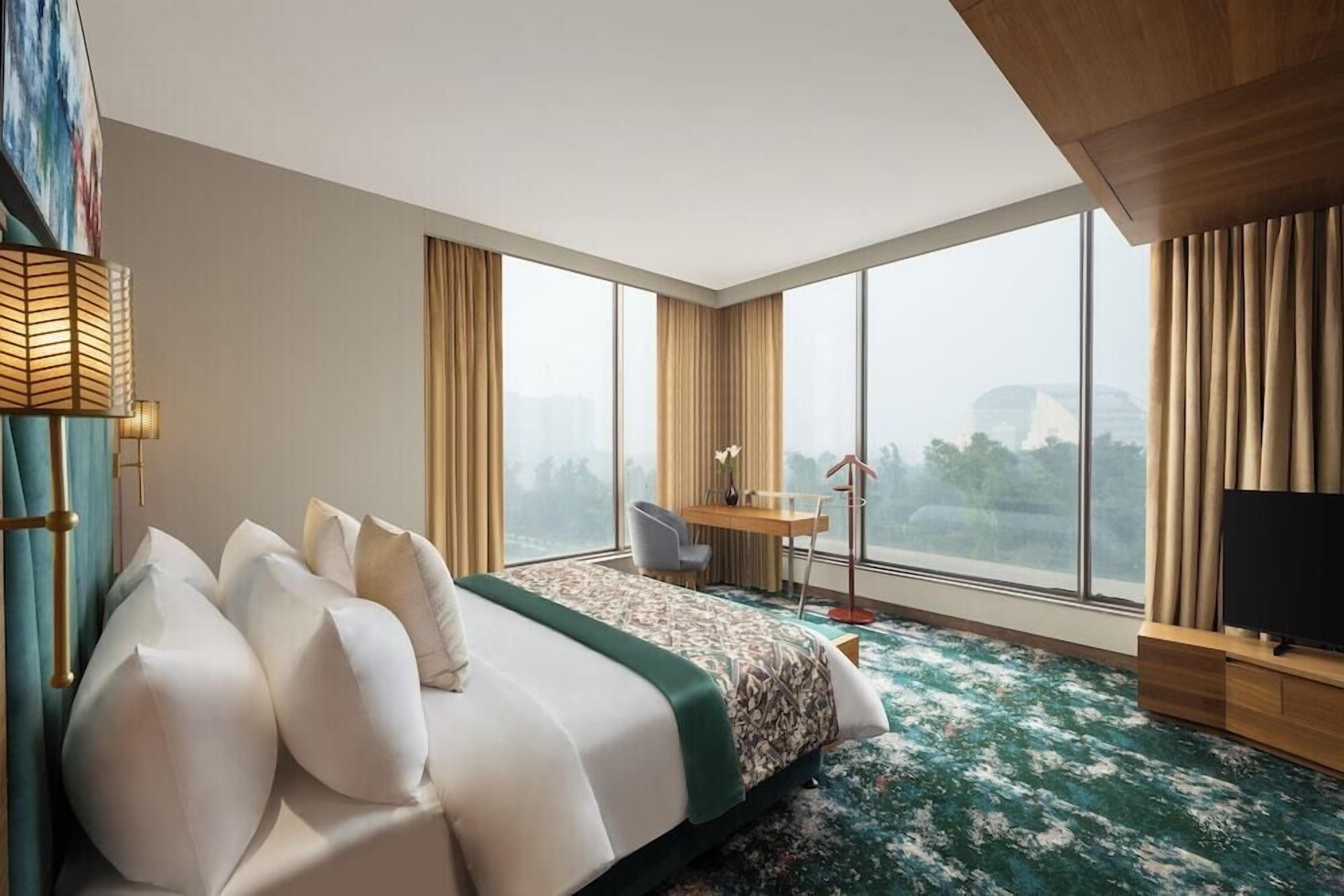 A hotel room with a large bed, a desk, a chair, a TV, and a view of the city through large windows.