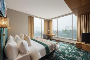 A hotel room with a large bed, a desk, a chair, a TV, and a view of the city through large windows. - Fortune Park East Delhi (New Delhi)