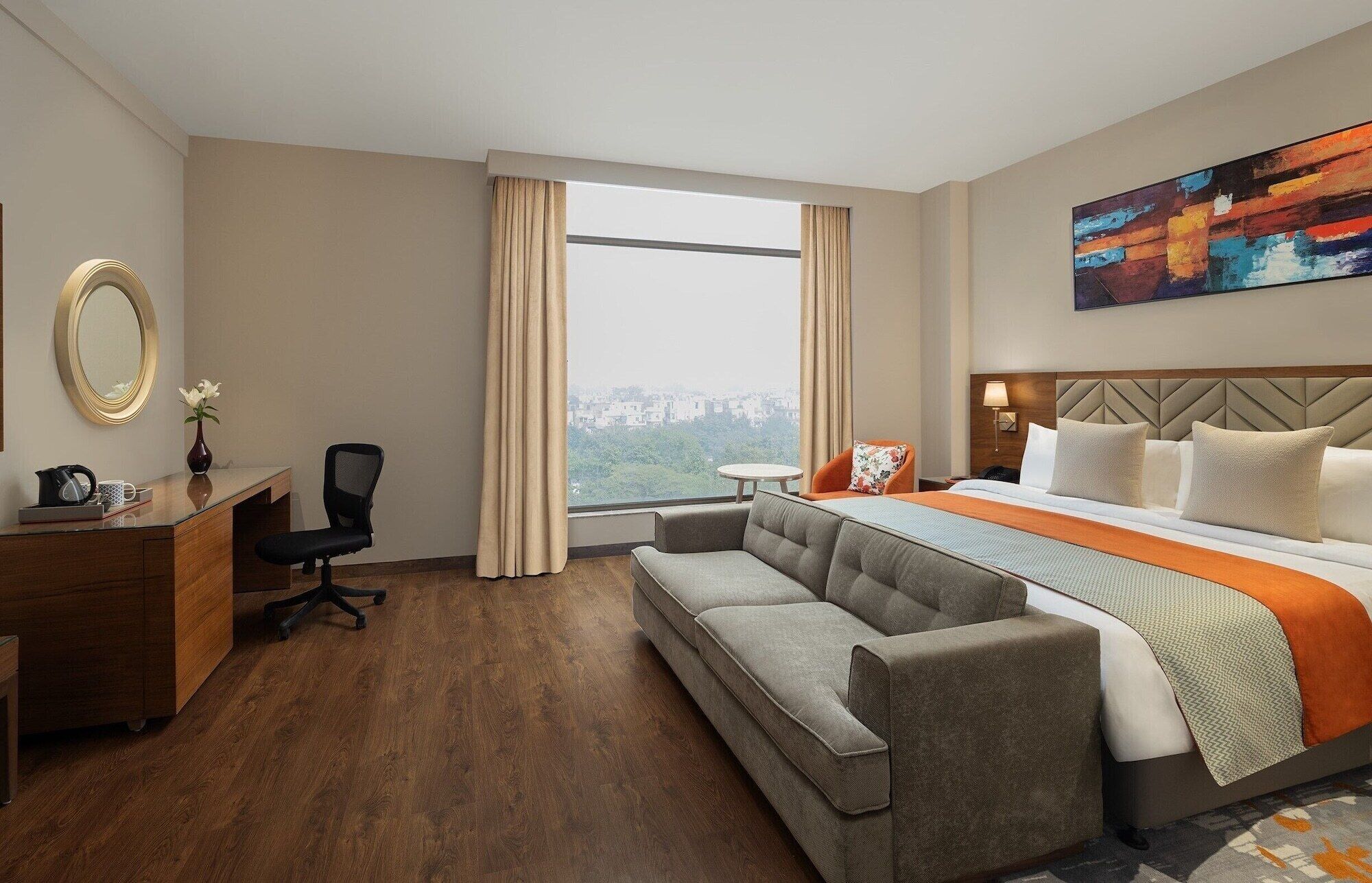 A hotel room with a bed, a sofa, a desk, a chair, a mirror, and a window with a city view.