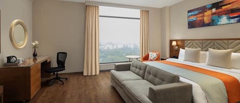 A hotel room with a bed, a sofa, a desk, a chair, a mirror, and a window with a city view.