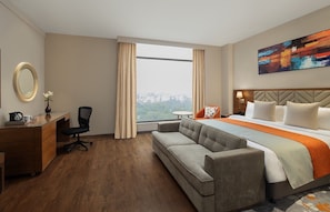 A hotel room with a bed, a sofa, a desk, a chair, a mirror, and a window with a city view. - Fortune Park East Delhi (New Delhi)