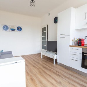 A modern kitchen with white cabinets, a built-in oven, and a microwave. There is a bench with a flat-screen TV, and a clock on the wall. - Apparthotel T3 Epicservices Saint-charles (Marseille)