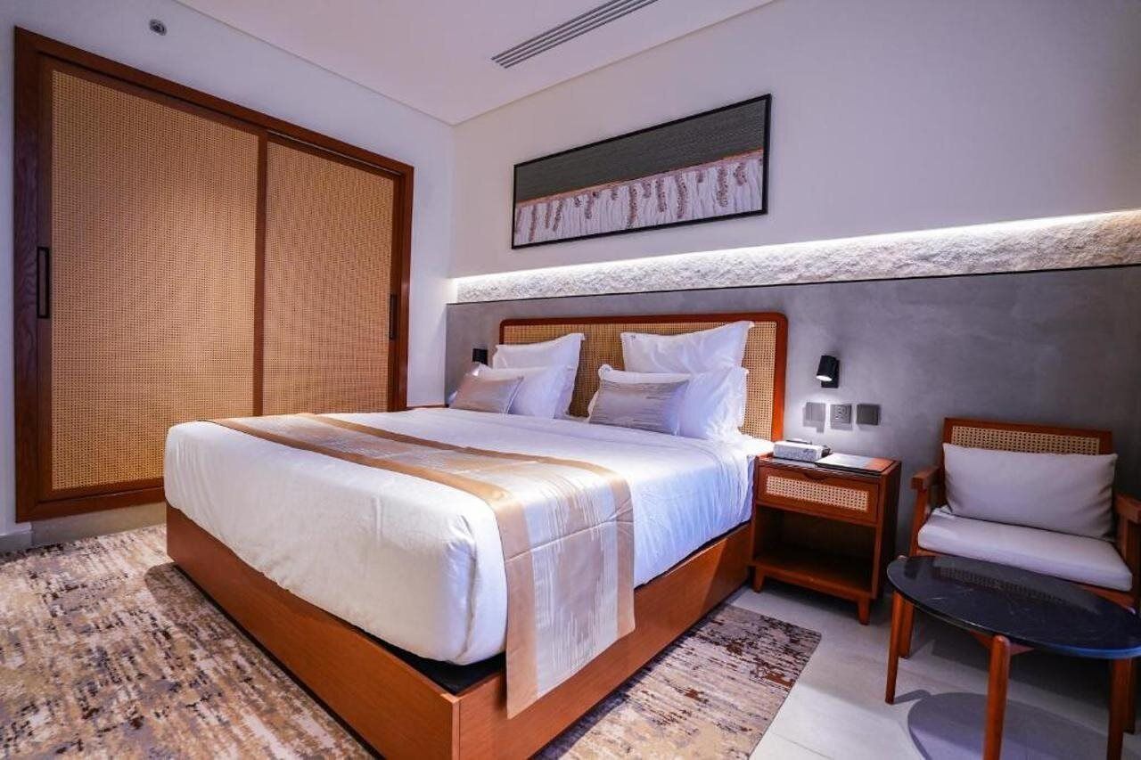A modern hotel room with a large bed, a wooden nightstand, a chair, and a large window.