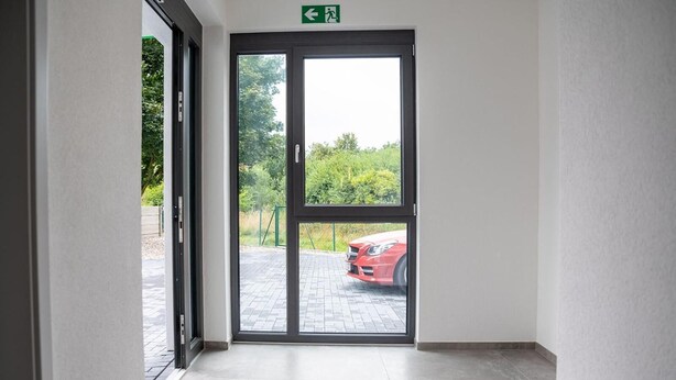 A hotel entrance with a glass door, a green exit sign, and a view of a paved area with a red car. - Hotel Grune Elster (Fuerth)