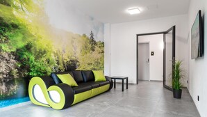 A modern lobby with a green and black sofa, a small table, a potted plant, and a mural of a forest scene. - Hotel Grune Elster (Fuerth)