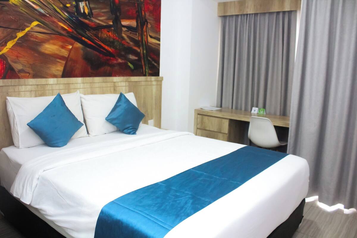 Ein Hotelzimmer mit einem Bett, blauen Kissen, einer blauen Bettdecke, einem Schreibtisch mit Stuhl und einem Bild an der Wand.