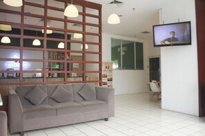 A modern interior with a grey sofa, pendant lights, a TV on the wall, and a reception area with a counter and menu board. - Urban Hotel Serpong by Zuzu (South Tangerang)