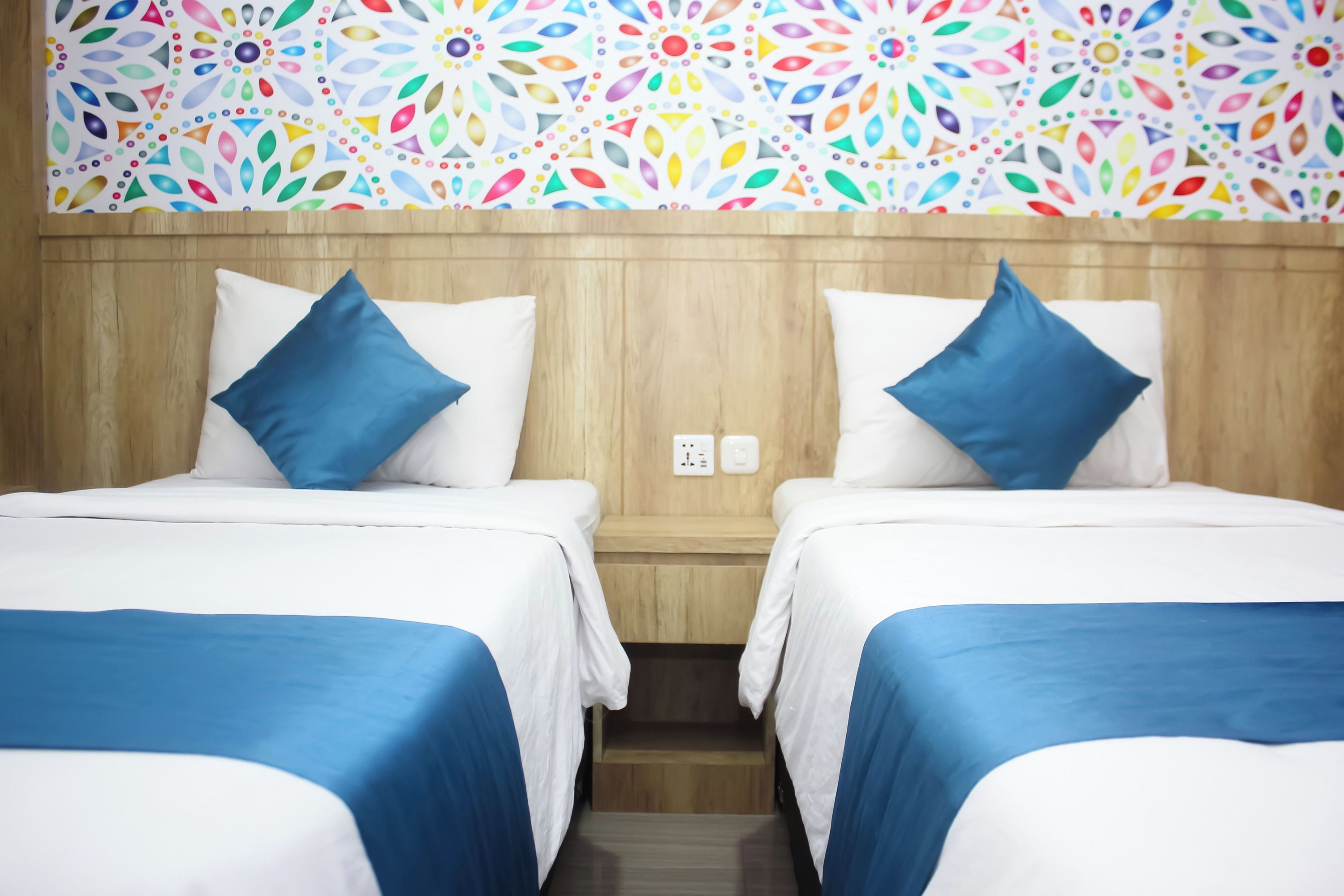 A hotel room with two beds, each with white bedding and blue accents, a wooden headboard, and a colorful floral wall pattern.