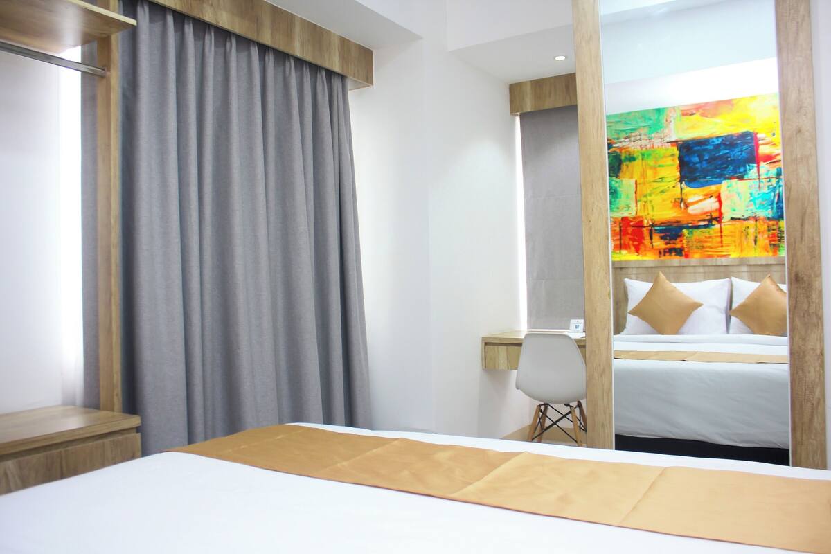 Ein Hotelzimmer mit einem Bett, einem Schreibtisch, einem Stuhl und einem großen Spiegel, der ein farbenfrohes abstraktes Gemälde widerspiegelt.