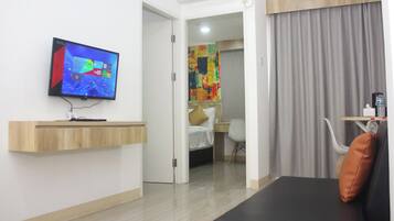 A hotel room with a flat-screen TV mounted on the wall, a wooden shelf, a bed with a white headboard, a desk with a chair, and a large window with curtains.