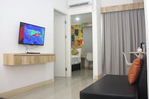 Camera d'albergo con televisione a schermo piatto montata a parete, una mensola in legno, un letto con testiera bianca, una scrivania con sedia e un'ampia finestra con tende.